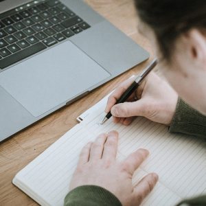 man writing in notebook next to computer